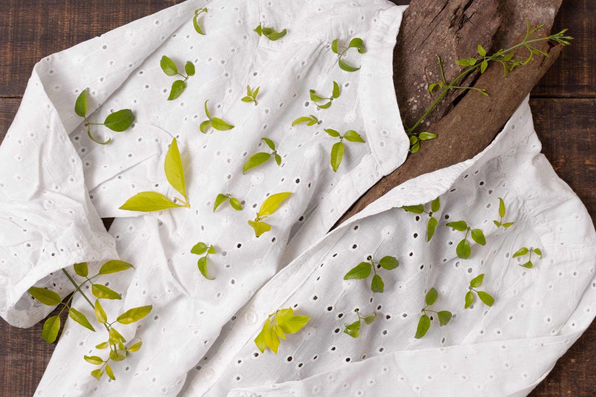 A white eyelet blouse laid on wood and scattered with fresh leaves.