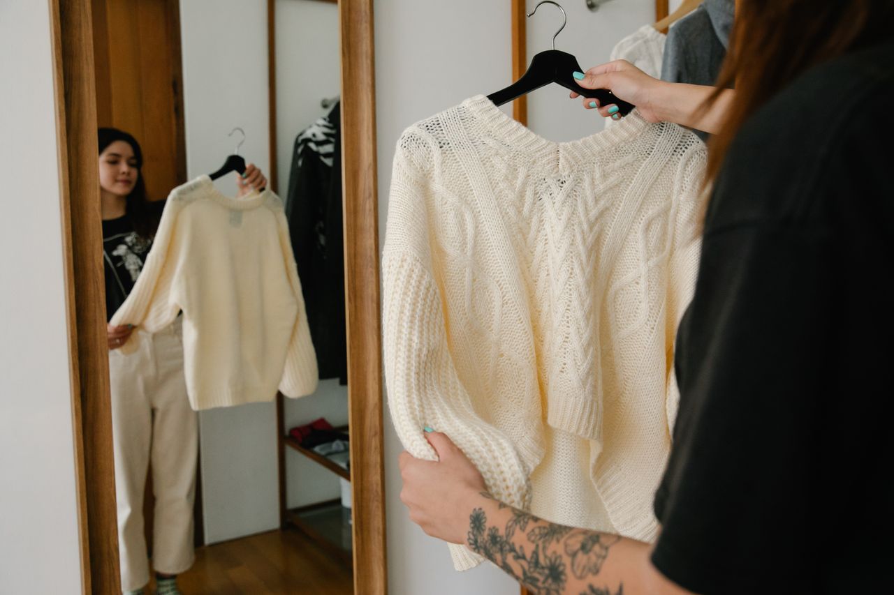 A cream cable-knit sweater checked on a hanger at a mirror.